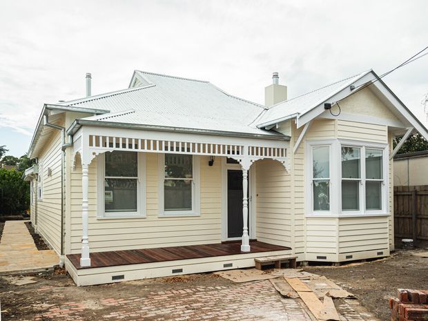 Exterior view of a renovated single-storey heritage-style weatherboard house with a pale cream facade, light grey corrugated metal pitched roof, ornate covered front veranda, decorative timber fretwork, bay window, and timber decking. The house sits on a paved and partially unfinished site with a timber boundary fence.