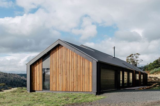 A compact contemporary gable-roofed house viewed from outside, set on a sloping rural site. The facade combines vertical warm timber boards with dark metal-clad walls and a dark pitched metal roof. Large glazed openings and a garage door run along the side, with a gravel driveway in the foreground and a wooded landscape beyond.