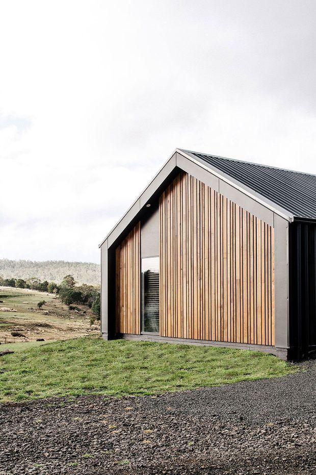 Exterior view of a compact contemporary rural building with a simple gabled form. The facade combines vertically arranged warm timber boards with dark metal framing and a ribbed dark metal pitched roof. A narrow glazed opening interrupts the timber facade, while grass, gravel, and rolling wooded countryside surround the structure.