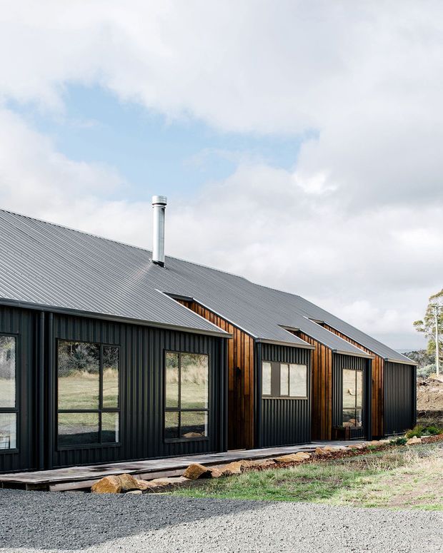 Exterior view of a long, low contemporary building with repeated gabled roof volumes, dark vertical corrugated metal cladding, black-framed glazing, and warm timber-clad recessed sections. A gravel driveway and sparse landscaped ground sit in front, with open rural terrain beyond. The image is dominated by the building facade and roofline under a cloudy sky.
