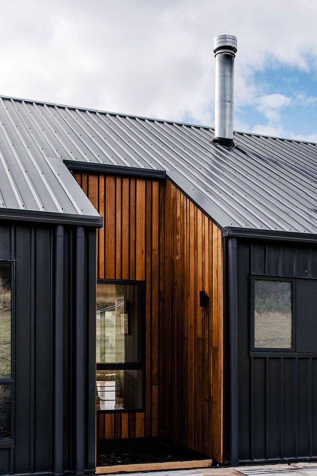 Close-up exterior view of a compact modern building with intersecting pitched roof forms. The facade combines dark vertical metal cladding with warm vertical timber weatherboarding, large dark-framed glazing, and a prominent cylindrical metal chimney. The image focuses on the roofline and entrance facade against an overcast sky and rural-looking surroundings.