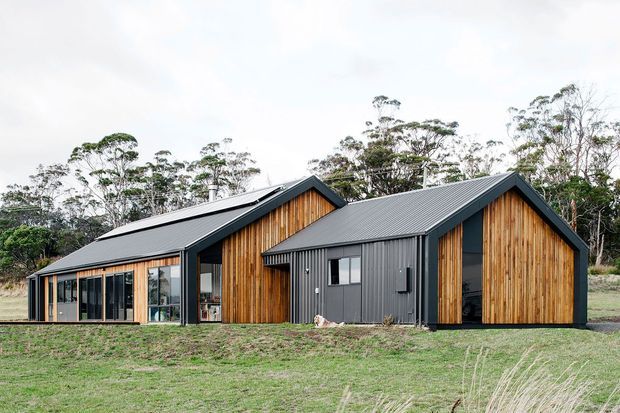 A contemporary single-storey residential building set in a rural, wooded landscape. The composition consists of connected gabled volumes with dark grey corrugated metal roofing and cladding, contrasted by warm vertical timber wall cladding. Large glazed openings are visible along the left wing, while the central dark-clad section includes a small window and a recessed entry or opening.