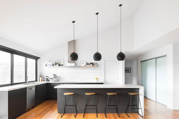 Bright contemporary kitchen with a large central island and four bar stools, dark lower cabinetry, white tiled splashback, white benchtops, timber flooring, black-framed glazing, and three black pendant lights. A sliding glazed door and hallway are visible beyond the kitchen.
