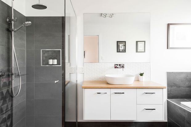 Bright contemporary bathroom featuring a walk-in glass shower with dark grey wall tiles, a white vessel basin on a light timber benchtop, white built-in vanity cabinetry, patterned white wall tiles, and a partially visible freestanding or built-in bath.