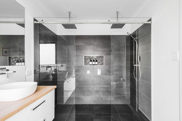 Contemporary bathroom featuring a large frameless glass walk-in shower with dual overhead rainfall showerheads and a handheld shower. Dark grey large-format tiled walls and floor create a strong contrast with the white floating vanity, oval vessel basin, and light timber benchtop. A mirrored wall and recessed shower niche add reflective and functional detail.
