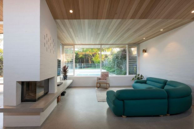 Bright contemporary living room with a large teal sectional sofa, a white masonry fireplace, pale concrete flooring, extensive garden-facing glazing, and a dramatic timber-lined ceiling with recessed lighting. A pool and landscaped garden are visible beyond the windows.