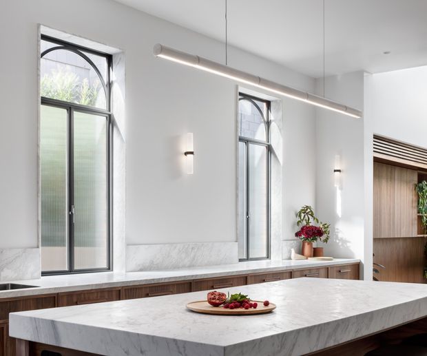 Bright, refined kitchen interior with a large white marble island in the foreground, dark timber cabinetry, white walls, tall arched-top black-framed windows, and a linear suspended light. A marble backsplash or ledge runs beneath the windows, with a timber-lined opening and indoor plants at the right.