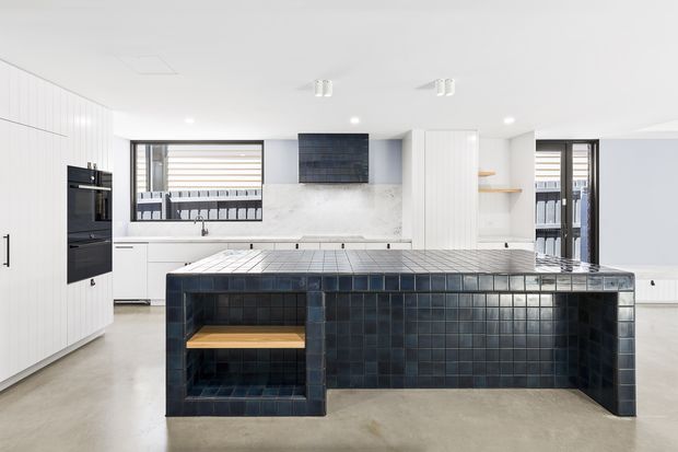 Bright contemporary kitchen featuring a large island clad in dark blue-black square tiles, with integrated open timber shelving. White vertical-panel cabinetry, pale stone or marble-look splashback, integrated black ovens, timber shelving, and generous windows create a clean, high-contrast space.