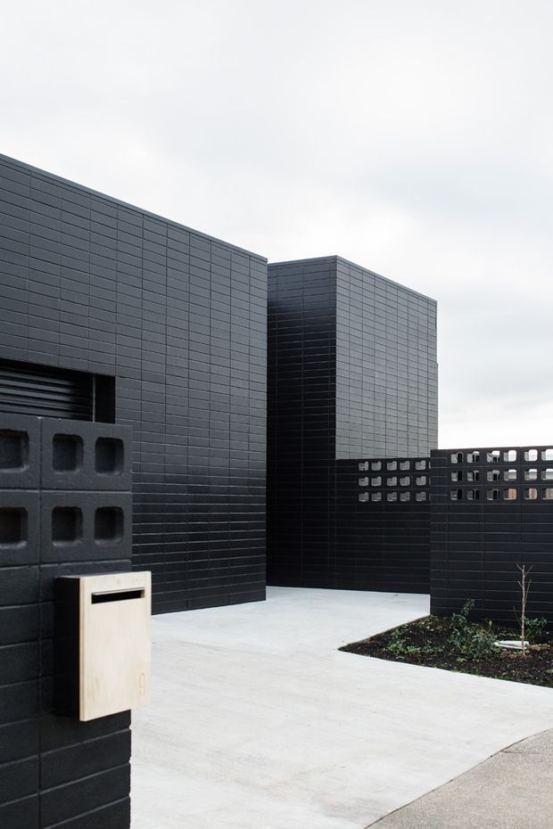 Exterior view of a contemporary, highly geometric property entrance. Dark charcoal masonry and finely gridded cladding form tall box-like volumes, with a perforated breeze-block boundary wall and pale concrete pathway. A minimalist timber-toned wall-mounted letterbox provides a small contrasting detail, while sparse planting sits beside the entry.