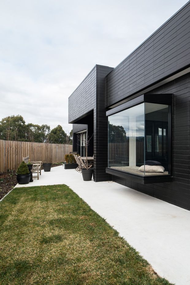 Exterior view of a contemporary single-storey home with strong geometric volumes, dark horizontal brick or masonry cladding, and extensive black-framed glazing. A projecting glass bay window overlooks a paved terrace, with outdoor seating, planters, timber fencing, and a small lawn in the landscaped side yard.