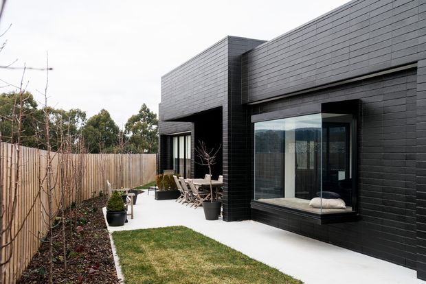A contemporary single-storey home exterior with a dark brick-clad facade, strong rectilinear volumes, and large glazed openings. A paved courtyard-style walkway leads past a lawn and planted garden bed to an outdoor dining area and sliding glass doors. Timber fencing and established trees frame the residential setting.