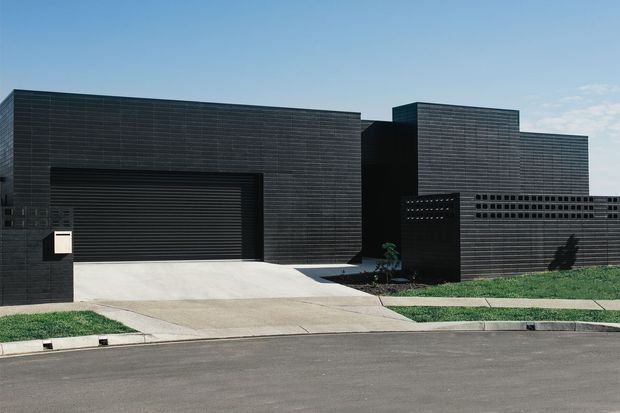 Street-facing view of a low, contemporary residential facade composed of dark horizontally coursed cladding, solid geometric volumes, a recessed entry, and a wide integrated garage door. The property sits behind a concrete driveway and simple lawn verge under clear daylight.