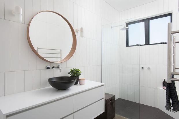 Bright contemporary bathroom featuring white vertical wall panelling, a white floating vanity with integrated drawers, a matte black vessel basin, round timber-framed mirror, black tapware, glass shower enclosure, and dark tiled flooring. A high horizontal window and simple wall sconces add natural light and a clean, refined feel.