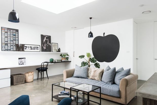 Bright, open-plan contemporary living room with a neutral sofa, black metal-and-glass coffee table, polished concrete flooring, white walls, built-in shelving, pendant lights, skylight, indoor plants, and a bold oversized apple wall graphic. A compact work area is integrated along the rear wall, and a kitchen or adjoining space is partially visible at right.