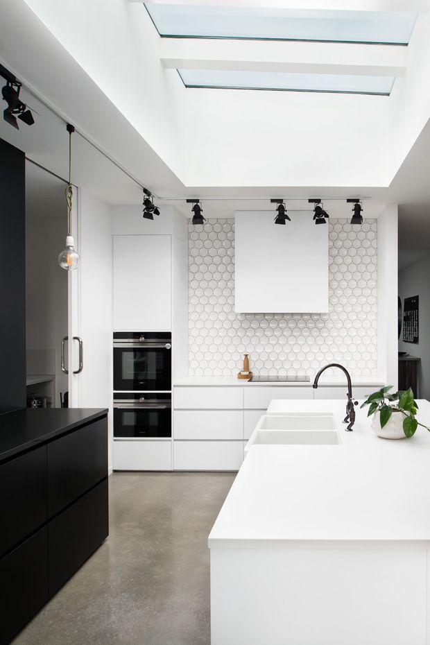 Bright contemporary kitchen with a large white island and integrated double sink, white flat-panel cabinetry, black appliances and dark island cabinetry. The rear wall features a repeating circular-pattern tile splashback and concealed rangehood, while exposed track lighting and large skylights bring strong illumination into the space. A black gooseneck tap, pendant light and potted plant add contrast.