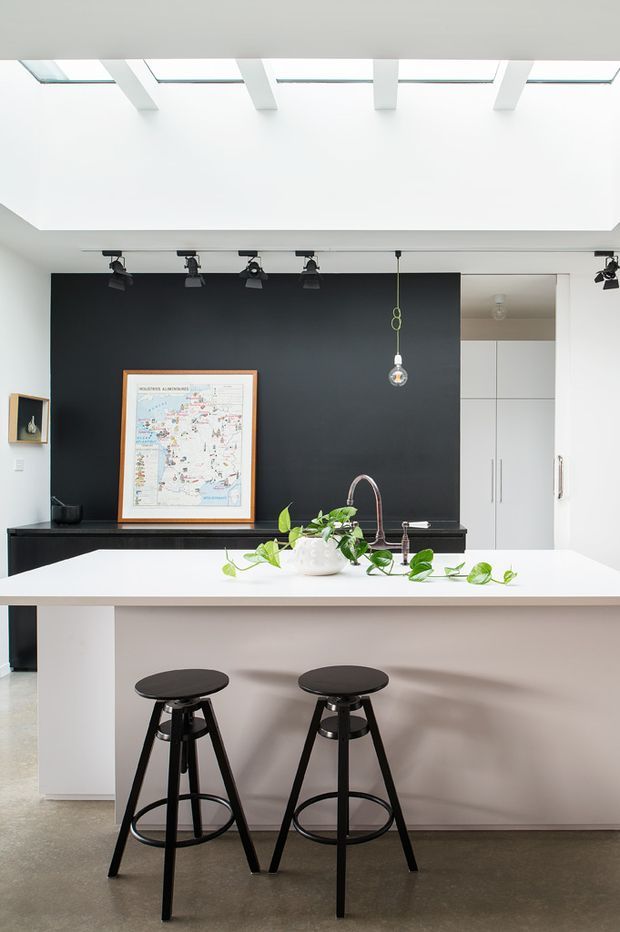 Bright contemporary kitchen featuring a large white island with seating for two, a white benchtop, integrated sink and curved faucet, dark cabinetry or backsplash wall, and black bar stools. A rooflight with white structural beams brings abundant natural light into the space, while track lighting and a pendant add layered illumination. White floor-to-ceiling cabinetry is visible at the rear, with a framed artwork and small display shelf against the dark wall.