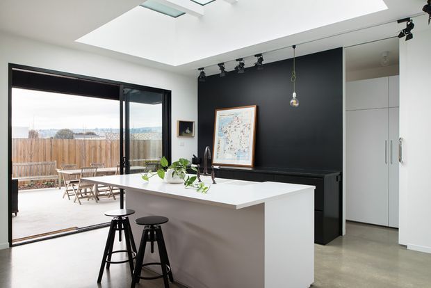 A bright contemporary kitchen with a large white island and two black bar stools, opening onto an outdoor terrace through wide glazed sliding doors. The room features a dark charcoal feature wall with track lighting, integrated white cabinetry, polished concrete-look flooring, a skylight, and a framed artwork.