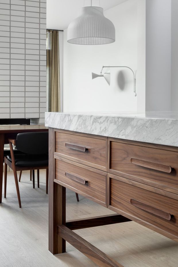 A close interior view of a refined kitchen and dining area. A walnut-toned timber island or sideboard with integrated drawers supports a thick white veined stone benchtop. Behind it, a black dining chair and timber table sit beside a white tiled wall, while a large ribbed pendant light and minimalist wall sconce add understated character. The space has a clean, warm, mid-century-inspired appearance with pale timber flooring and neutral white surfaces.