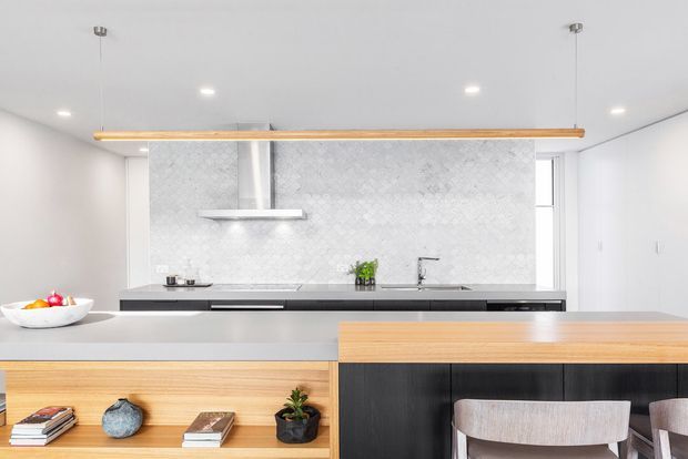 A bright, contemporary open-plan kitchen and dining area. The kitchen features a long dark island with a pale benchtop, light timber open shelving, dark lower cabinetry, a pale patterned tile splashback, integrated stainless-steel appliances, and a linear suspended timber pendant. A small dining area with light upholstered chairs is visible in the foreground, along with minimal decorative objects and recessed ceiling lighting.