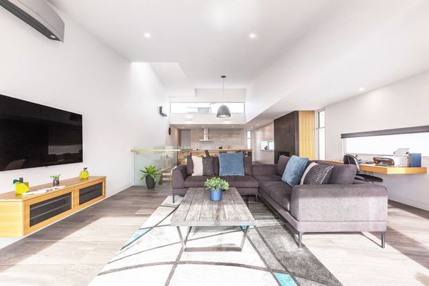 Bright open-plan contemporary living room with a large L-shaped grey sectional sofa, textured geometric rug, timber-framed coffee table, wall-mounted television and low timber media cabinetry. The living area opens toward a compact kitchen and dining zone, with white walls, recessed lighting, timber and dark joinery, pendant lighting, and extensive natural light from windows and clerestory glazing.