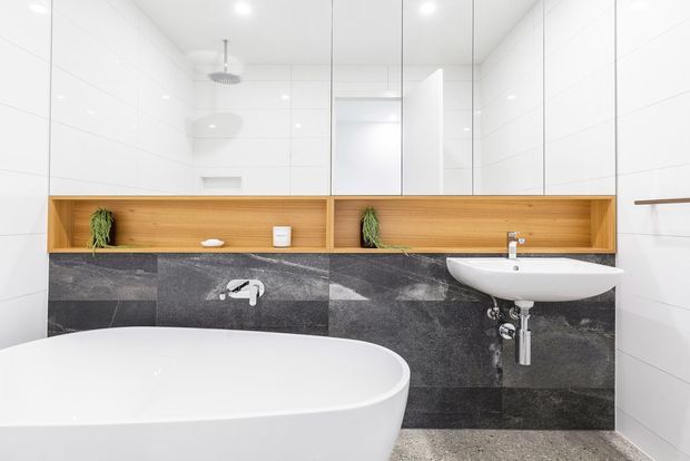 Bright contemporary bathroom featuring a freestanding white bathtub in the foreground, a wall-mounted white basin, large mirrored cabinetry, white wall tiles, and a dark stone-look tiled feature wall. A long recessed timber display shelf with integrated planter niches runs above the basin and bath, with chrome tapware and fittings.