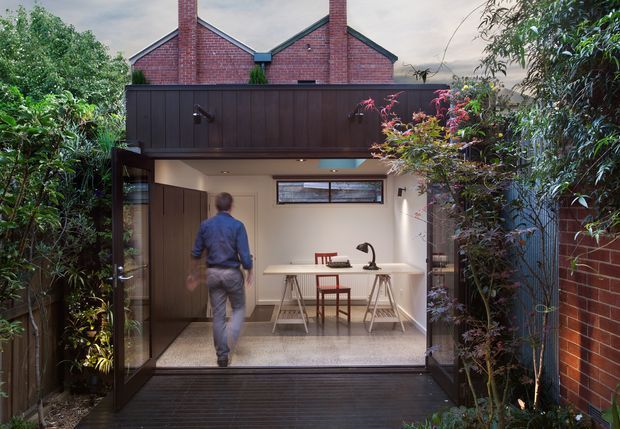 A compact contemporary kitchen extension opening onto a timber deck and enclosed garden courtyard. The kitchen has pale cabinetry and benchtops, a dark timber-clad overhead volume, integrated appliances, bar seating, and a long horizontal clerestory window. Behind it is an older red-brick gabled building, creating a clear contrast between heritage masonry and the modern addition.
