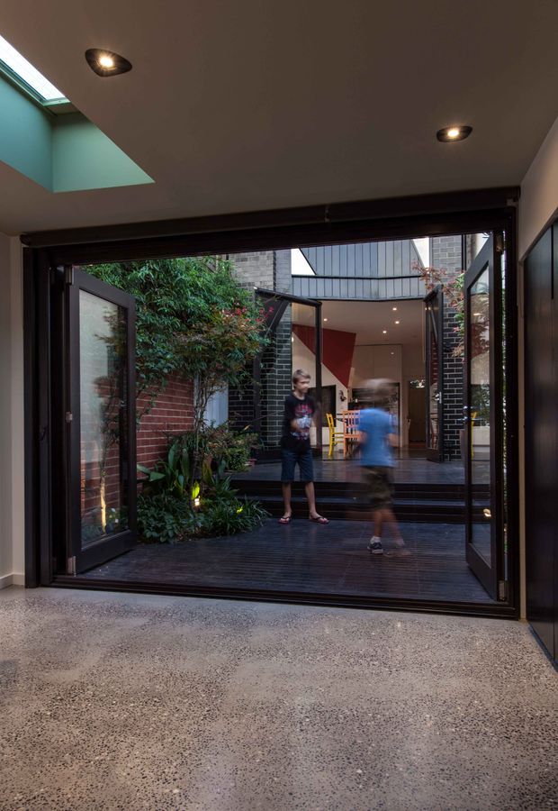 View from an interior space through fully opened dark-framed glazed doors onto a timber courtyard deck and planted courtyard. The contemporary dwelling features a polished exposed-aggregate concrete floor, dark window and door frames, brick courtyard walls, a skylight, recessed ceiling lights, and a connected interior beyond with glimpses of colourful furnishings and a high glazed roof or clerestory.