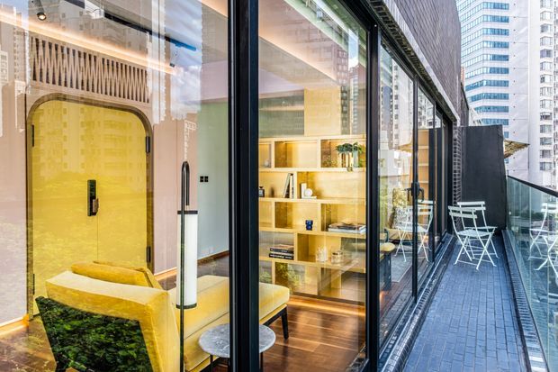 A high-rise apartment interior viewed through floor-to-ceiling glazing, showing a compact contemporary living area with a bright yellow upholstered armchair, dark green marble-like coffee table, timber shelving and cabinetry, and a large rounded yellow feature door or wall panel. A narrow balcony with dark tiled flooring, outdoor café furniture, and urban towers is visible to the right.