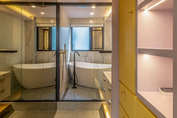 A refined contemporary bathroom viewed from the entry, featuring a freestanding oval bathtub, glass-enclosed walk-in shower, twin dark-framed windows, marble-look wall surfaces, warm integrated lighting, and built-in vanity and shelving. A partially visible toilet is located within illuminated built-in cabinetry at right.