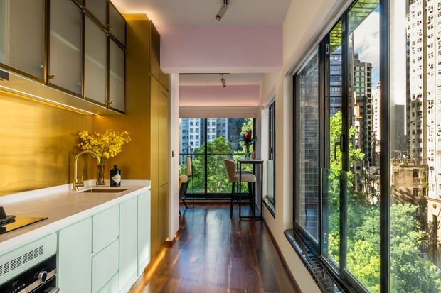 A compact contemporary kitchen opening into a narrow dining or living corridor, with pale mint cabinetry, a warm metallic gold backsplash, integrated upper storage, white benchtop, and timber flooring. Full-height black-framed glazing runs along the right side, overlooking a dense urban high-rise setting. A small dining table and chairs sit at the far end near a balcony or window.