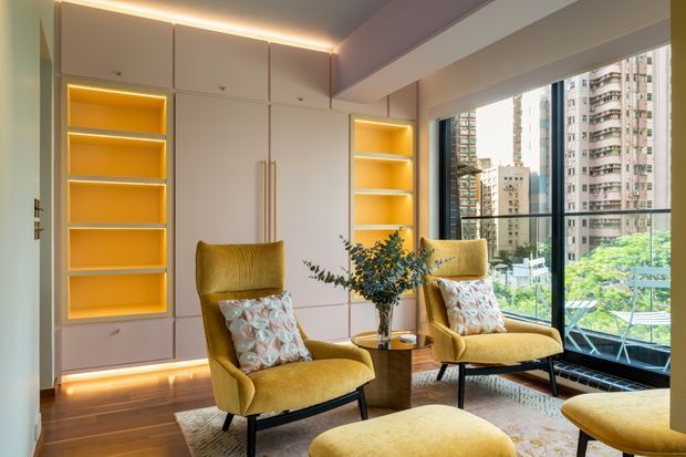A refined living room with custom pale cabinetry, illuminated recessed display shelving, warm integrated LED lighting, two mustard upholstered lounge chairs, a matching ottoman, patterned cushions, a small round side table, and a large floor-to-ceiling window overlooking an urban high-rise setting.