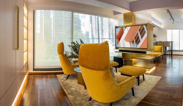 A stylish open-plan living room with a pair of sculptural mustard-yellow lounge chairs arranged around a television and low built-in media console. The room features dark timber flooring, a large neutral rug, floor-to-ceiling windows with roller blinds, integrated warm LED lighting, and a dining area visible in the background. Contemporary urban high-rise views are visible through the glazing.