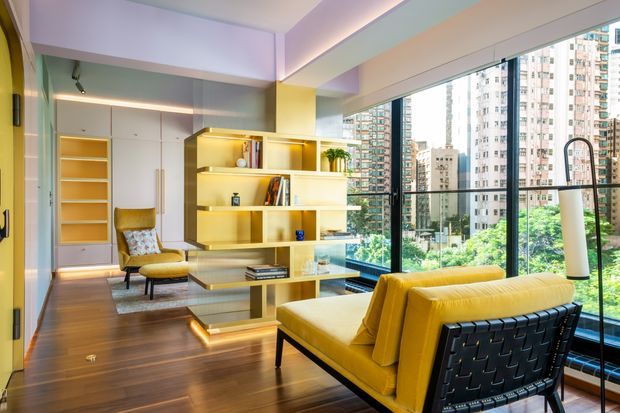 Bright contemporary living room in a high-rise apartment, featuring floor-to-ceiling glazing, a yellow upholstered sofa, mid-century-inspired lounge chair, warm timber flooring, and a built-in open shelving and media unit with integrated lighting. Dense urban apartment towers are visible outside.
