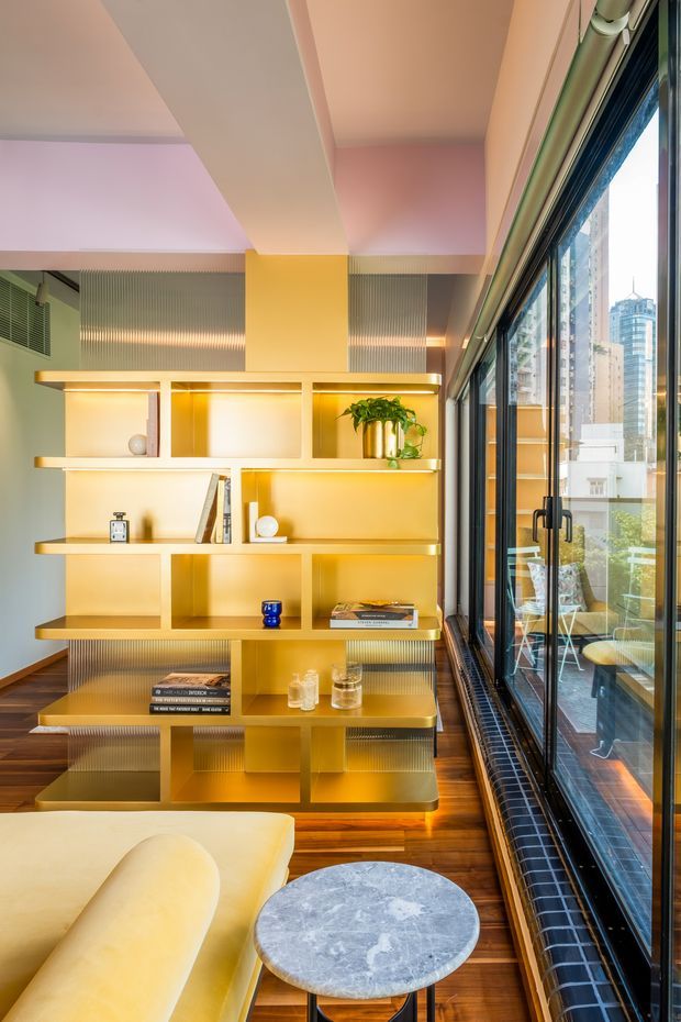 A stylish living room with a warm yellow built-in open shelving unit, integrated lighting, and ribbed translucent panels. The room features a curved cream sofa, round grey stone coffee table, rich timber flooring, and full-height black-framed glazing overlooking an urban cityscape. The ceiling and walls are softly tinted in blush and peach tones, creating a bright, refined contemporary interior.