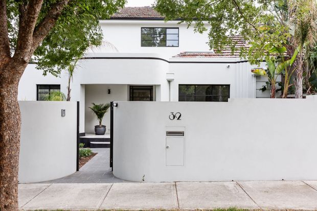 Street-facing view of a white rendered multi-level residence behind a curved boundary wall, with a pedestrian entry gate, concealed front porch, dark-framed windows, tiled pitched roof sections, and mature tropical planting.