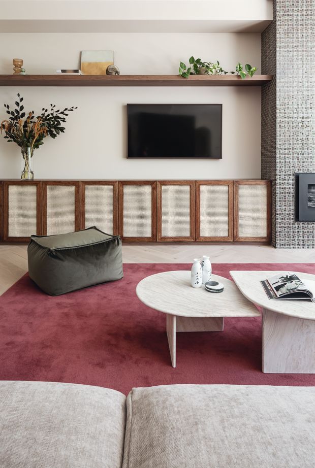 A refined living room centered on a wall-mounted television, with a long dark timber built-in media console featuring woven cane-front doors. Open timber shelving displays artwork and trailing plants, while a textured mosaic-tile feature wall frames the right side. The room includes a deep burgundy area rug, sculptural light wood coffee tables, a soft grey lounge chair, and a large neutral sofa in the foreground.