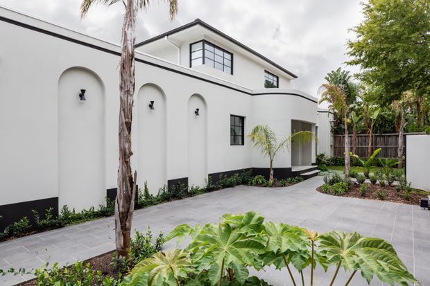 Contemporary white rendered residential exterior surrounding a landscaped courtyard. The facade features repeated tall arched recessed niches with small black wall lights, black base trim, dark-framed windows, and a simple pitched roof. Grey paved paths and planting beds with palms and tropical foliage create a lush courtyard setting.