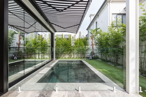 A contemporary enclosed courtyard pool area with a narrow rectangular swimming pool, pale stone paving, extensive floor-to-ceiling glass walls, and a black slatted pergola overhead. Lush bamboo and other planting line the boundaries, with adjacent white rendered multi-storey buildings visible beyond. The reflective glazing and dark pergola create strong linear patterns, while the pool and greenery give the space a calm, private atmosphere.