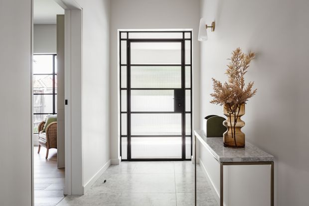 A refined residential entry hall with a black-framed glazed front door featuring translucent ribbed glass, pale tiled flooring, white walls and trim, and a slim stone-topped console with integrated cabinetry. A wall sconce and dried botanical arrangement add warmth. An adjacent furnished living area is visible through the open doorway on the left.