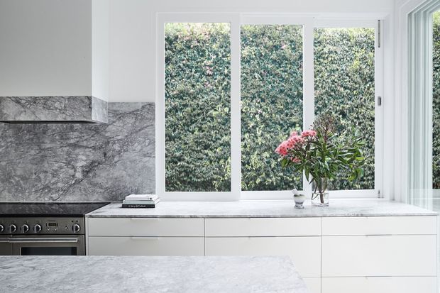 Bright contemporary kitchen with extensive white built-in cabinetry, a pale marble island and benchtop, and a dramatic grey-veined marble splashback. Large white-framed windows overlook dense greenery, while a freestanding vase with flowering branches adds a natural accent.