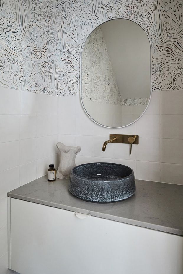 A compact bathroom vanity with a round dark textured vessel basin set on a grey stone benchtop. White tiled walls are paired with distinctive monochrome patterned wallpaper above, while a large rounded-rectangle mirror and brushed brass wall-mounted tap add decorative detail. White handleless cabinetry sits below the vanity.