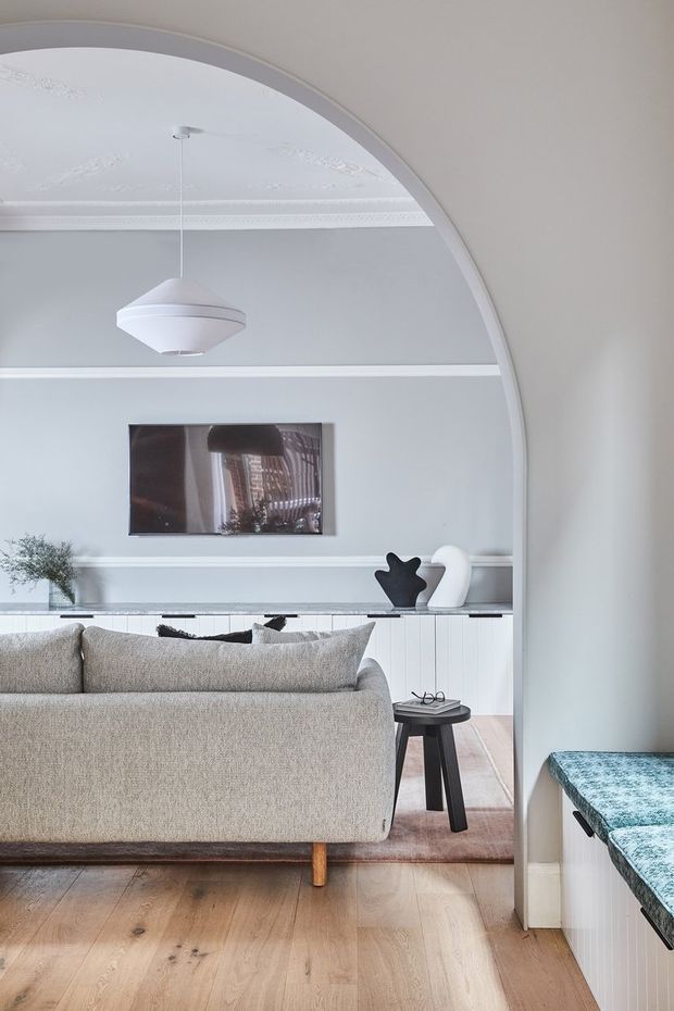 A refined living room viewed through a large arched opening, featuring a light neutral sofa, pale timber flooring, white built-in cabinetry, minimalist sculptural decor, and a suspended geometric pendant light. Decorative ceiling mouldings and a curved architectural arch add character, while a blue-green stone or patterned benchtop is visible in the foreground.