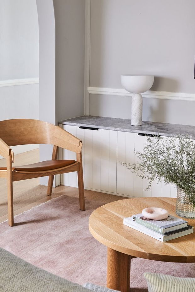 A softly styled living room corner featuring a sculptural mid-century wooden armchair, a round timber coffee table, and a low built-in white panelled cabinet with a grey stone-look benchtop. The room has pale taupe walls, decorative wall moulding, a pink-toned rug, a ceramic table lamp, greenery, and layered neutral furnishings. The overall look is warm, refined, and contemporary with mid-century influences.