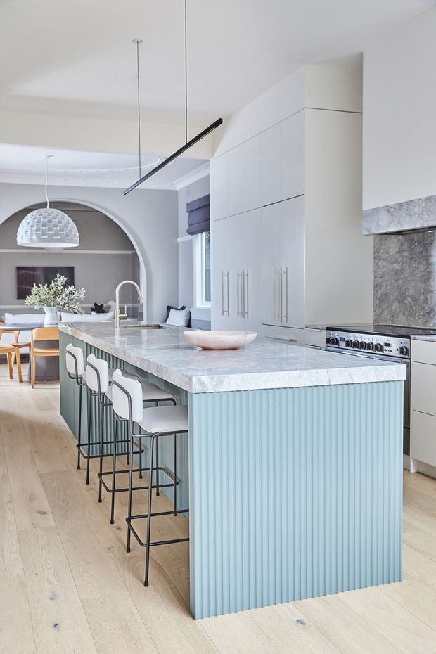 Bright, refined kitchen interior with a large marble-look island featuring a pale blue fluted island base and seating for several people. The kitchen includes tall white cabinetry with long vertical handles, a matching stone benchtop and splashback, a stainless steel range cooker, pale timber flooring, and contemporary black linear pendant lighting. An arched opening leads to a dining or living area with classic decorative detailing.