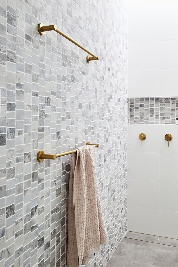 A bright contemporary bathroom with grey and white mosaic tile walls, large light grey floor tiles, and brushed brass shower and towel rail hardware. A pale textured towel hangs from the lower rail, while a contrasting white tiled wall with a horizontal mosaic accent and brass controls is visible in the shower area.