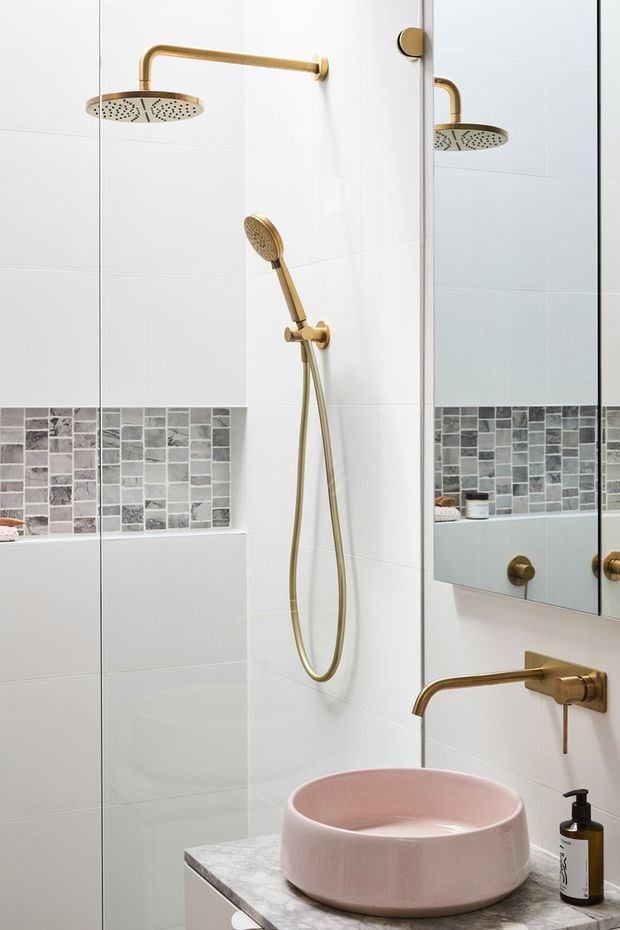 Contemporary bathroom featuring a frameless glass shower enclosure, large white wall tiles, a horizontal band of grey mosaic accent tiles, and brushed brass shower and basin fittings. A round blush-pink vessel basin sits on a marble-look vanity, with a large mirror reflecting the shower.