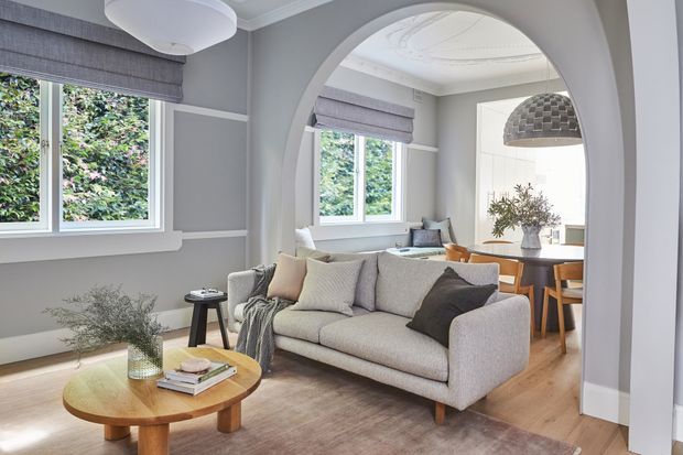 A refined open-plan living and dining area viewed through a large arched opening. The room features a light upholstered sofa, round timber coffee table, timber flooring, grey painted walls with decorative panel moulding, large windows with Roman blinds, and a dining table beneath a woven pendant light. The palette is soft grey, white, beige, and natural timber, creating a bright, calm and inviting atmosphere.