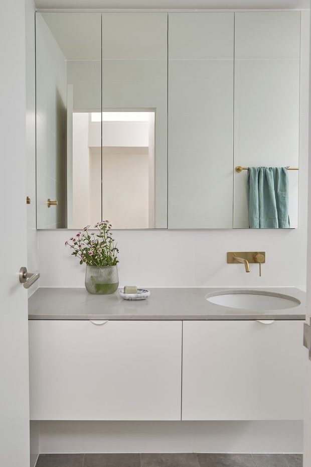 A bright, compact contemporary bathroom vanity with a wall-to-wall mirrored cabinet, pale grey stone benchtop, integrated oval basin, white floating cabinetry, and brushed brass wall-mounted tapware. A muted blue-green towel and small vase provide restrained accents.
