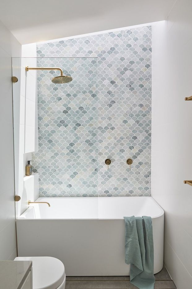 A compact contemporary bathroom featuring a white built-in bathtub enclosed by clear glass, a full-height scalloped mosaic tile feature wall in pale blue, grey and white, and brushed brass shower and tapware. White walls and sanitaryware create a clean, bright look, with a soft blue towel adding a subtle accent.