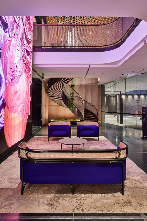 A dramatic upscale commercial lobby or hotel reception lounge featuring deep purple upholstered seating, a round coffee table, a large pink floral digital artwork or illuminated wall feature, polished dark flooring, and a sweeping sculptural staircase. The space has a curved mezzanine with a decorative patterned metal or timber balustrade, glass partitions, warm timber wall finishes, and layered contemporary lighting.